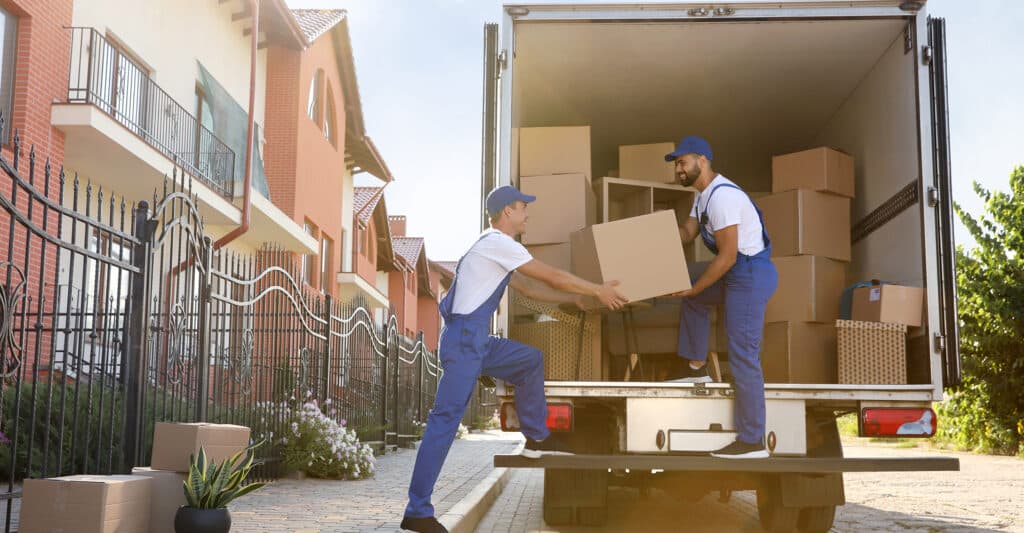 Deux déménageurs souriants en uniforme bleu chargent des cartons dans un camion de déménagement devant des maisons résidentielles.