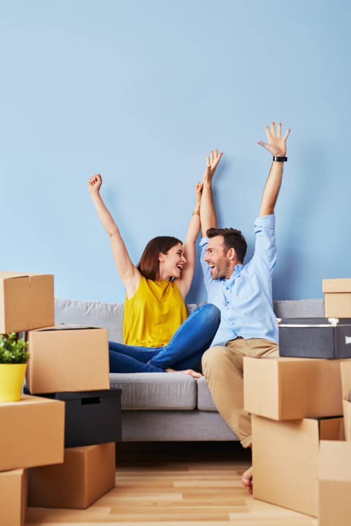 Couple ravi célèbre le déménagement, entouré de cartons dans leur nouveau logement au mur bleu. Bras levés de joie.