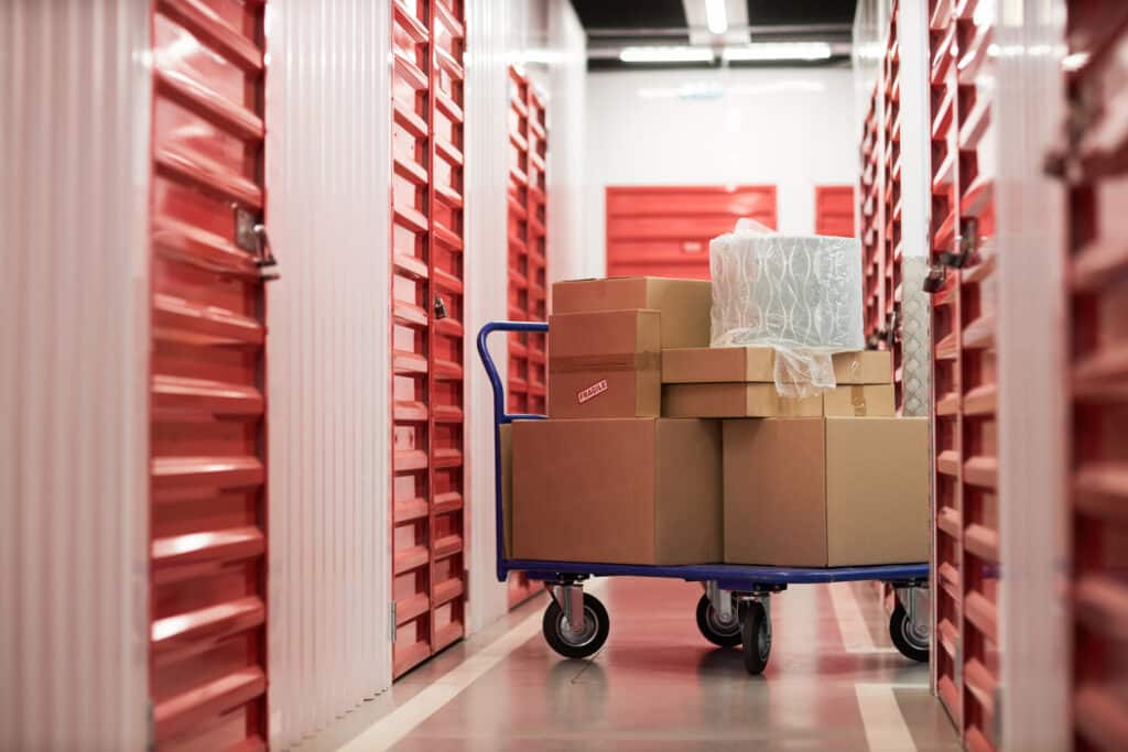 Chariot bleu rempli de boîtes en carton "FRAGILE" dans un couloir d'entrepôt avec unités de stockage rouges.